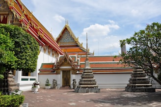 A traditional Thai temple with ornate, decorative roofs and gables highlighted by gold embellishments. The complex exhibits vibrant colors with intricately designed stupas in the foreground set on a paved courtyard. Lush greenery surrounds the temple, complementing the vibrant architecture, under a clear blue sky with scattered clouds.