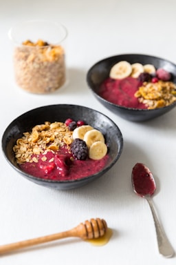 Two black bowls are filled with a vibrant berry smoothie base, topped with banana slices, blackberries, granola, and a sprinkle of red berries. A glass container filled with granola is in the background, and a honey dipper rests on the table next to a spoon with berry remnants.