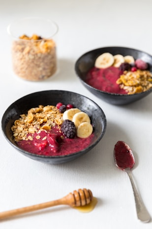 Two black bowls are filled with a vibrant berry smoothie base, topped with banana slices, blackberries, granola, and a sprinkle of red berries. A glass container filled with granola is in the background, and a honey dipper rests on the table next to a spoon with berry remnants.
