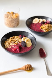 Two black bowls are filled with a vibrant berry smoothie base, topped with banana slices, blackberries, granola, and a sprinkle of red berries. A glass container filled with granola is in the background, and a honey dipper rests on the table next to a spoon with berry remnants.