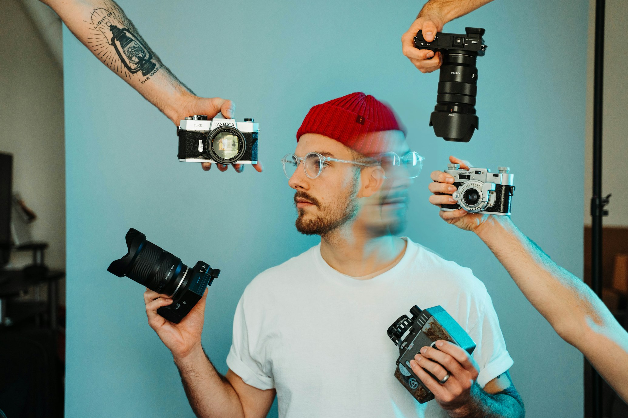 Man wearing white shirt and red hat holds two cameras while three other hands hold cameras near his face.