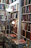 A cozy and densely packed room full of bookshelves lined with a wide variety of books. Piles of magazines and comics are stacked on a table in the foreground. A floor lamp with two glowing bulbs illuminates the space, creating a warm and inviting atmosphere.
