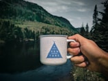 A hand is holding a white speckled mug with a blue triangle design and the word 'ADVENTURE' printed on it. The background features a scenic landscape with a river, dense green forests, and mountains under a cloudy sky.