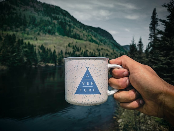 A hand is holding a white speckled mug with a blue triangle design and the word 'ADVENTURE' printed on it. The background features a scenic landscape with a river, dense green forests, and mountains under a cloudy sky.