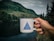 A hand is holding a white speckled mug with a blue triangle design and the word 'ADVENTURE' printed on it. The background features a scenic landscape with a river, dense green forests, and mountains under a cloudy sky.