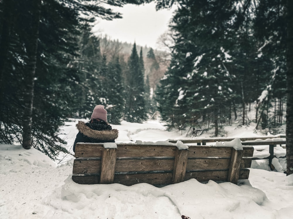 pessoa em jaqueta marrom sentada no banco de madeira marrom no chão coberto de neve durante o dia