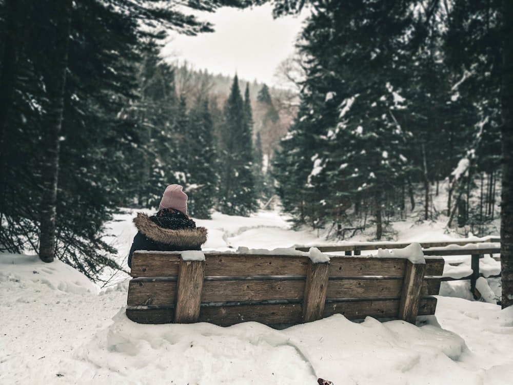 personne en veste brune assise sur un banc en bois brun sur un sol enneigé pendant la journée