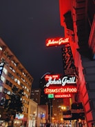 A vibrant city street at night features brightly lit signs for John's Grill, promoting jazz nights, steaks, seafood, and cocktails. The signs are illuminated in neon colors, casting a warm glow on the ornate building facades. Modern buildings and trees line the street, with additional neon lighting in the background enhancing the urban ambiance.