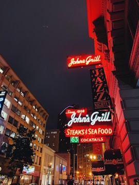 A vibrant city street at night features brightly lit signs for John's Grill, promoting jazz nights, steaks, seafood, and cocktails. The signs are illuminated in neon colors, casting a warm glow on the ornate building facades. Modern buildings and trees line the street, with additional neon lighting in the background enhancing the urban ambiance.