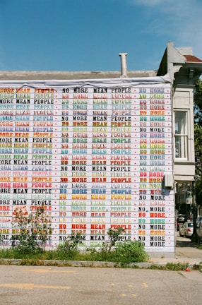 The side of a building is covered with repeating text that says 'NO MORE MEAN PEOPLE' in various colors and fonts. The wall is adorned with graffiti-style writing and small red heart icons. Greenery grows at the base of the building, and the sky is clear and blue.