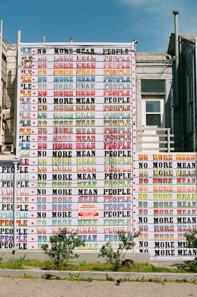 A large mural on a building exterior features the repeated phrase 'No More Mean People' in colorful, bold letters. The mural displays a variety of colors, creating a vibrant and eye-catching pattern. In front of the building, small plants and a gravel path are visible.