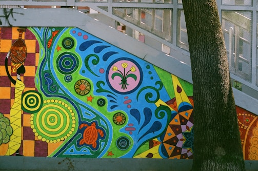 A vibrant wall mural featuring intricate patterns and symbols. The mural includes colorful geometric shapes, floral and animal motifs, such as a turtle and a stylized plant with flowers. The background showcases a mix of swirling lines and dots, creating a dynamic and lively composition. A tree trunk partially obscures the right side of the mural.