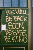 Large handwritten text in yellow spray paint reads 'WE WILL BE BACK SOON, BE SAFE, BE KIND' on a green boarded-up window. A small paper notice is taped to the bottom left corner.