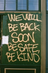 Large handwritten text in yellow spray paint reads 'WE WILL BE BACK SOON, BE SAFE, BE KIND' on a green boarded-up window. A small paper notice is taped to the bottom left corner.