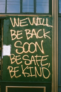 Large handwritten text in yellow spray paint reads 'WE WILL BE BACK SOON, BE SAFE, BE KIND' on a green boarded-up window. A small paper notice is taped to the bottom left corner.
