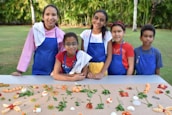 A group of five children wearing colorful aprons and towels over their shoulders stand behind a table arranged with an assortment of fruits and vegetables. They are outdoors in a garden setting with lush green trees in the background.