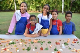 A group of five children wearing colorful aprons and towels over their shoulders stand behind a table arranged with an assortment of fruits and vegetables. They are outdoors in a garden setting with lush green trees in the background.