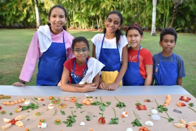 A group of five children wearing colorful aprons and towels over their shoulders stand behind a table arranged with an assortment of fruits and vegetables. They are outdoors in a garden setting with lush green trees in the background.