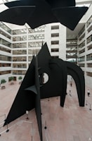 A large black metal abstract sculpture dominates the center of a spacious, modern atrium with multiple levels and glass-paneled walls. The sculpture features geometric shapes, sharp angles, and curves, and is surrounded by barriers. The atrium has light-colored stone floors and potted trees scattered throughout. Natural light floods in from the ceiling above.