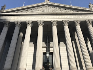 A grand neoclassical facade with tall, white Corinthian columns supporting a triangular pediment. Sculptures and inscriptions are visible above the columns, suggesting an official or governmental purpose.