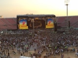 A large stadium filled with a crowd gathered in front of a stage. The stage features two large screens displaying the iconic Rolling Stones tongue logo and the word 'Santiago'. The atmosphere suggests a concert setting, with many people standing in close proximity and large speakers visible on either side of the stage.