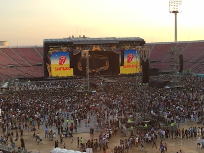 A large stadium filled with a crowd gathered in front of a stage. The stage features two large screens displaying the iconic Rolling Stones tongue logo and the word 'Santiago'. The atmosphere suggests a concert setting, with many people standing in close proximity and large speakers visible on either side of the stage.
