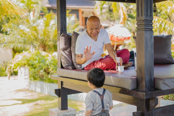 An adult is sitting on a shaded outdoor platform, wearing a white shirt and red pants, holding a mug. The setting appears tropical with sunlight filtering through palm leaves, casting shadows on the ground. A small child stands facing the adult, wearing a light-colored outfit with suspenders. The scene conveys a serene and relaxed atmosphere with lush greenery surrounding the area.