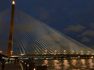 Nighttime shot of a luminous cable-stayed bridge lit up, reflecting on the river surface.