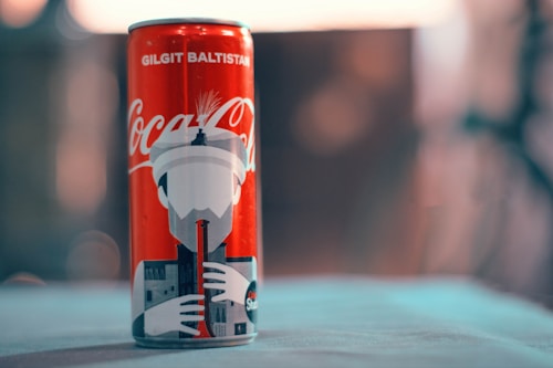 A Coca-Cola can with a design featuring the region Gilgit Baltistan. It includes an illustration of a person playing a traditional musical instrument against a backdrop of architectural elements.