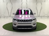 A silver SUV is parked on a small circular patch of artificial grass against a marble-patterned wall in a showroom. The car's design features distinct front grilles, headlights, and a reflection of pink and white lighting on the windshield.