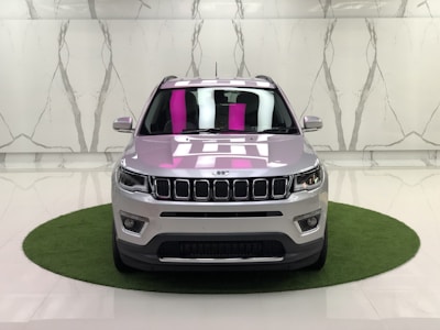 A silver SUV is parked on a small circular patch of artificial grass against a marble-patterned wall in a showroom. The car's design features distinct front grilles, headlights, and a reflection of pink and white lighting on the windshield.