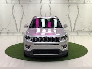 A silver SUV is parked on a small circular patch of artificial grass against a marble-patterned wall in a showroom. The car's design features distinct front grilles, headlights, and a reflection of pink and white lighting on the windshield.