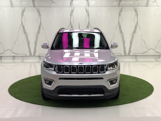 A silver SUV is parked on a small circular patch of artificial grass against a marble-patterned wall in a showroom. The car's design features distinct front grilles, headlights, and a reflection of pink and white lighting on the windshield.