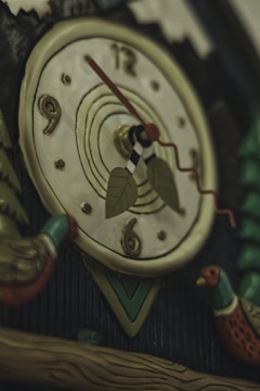 A close-up view of a decorative clock face featuring stylized numbers, with hands pointing to approximately 9:15. The clock is adorned with leaf-shaped pendulum weights and appears to be part of a traditional cuckoo clock design, with colorful bird figures and wooden elements.