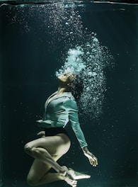A person underwater, elegantly posed with knees bent and head tilted back. Bubbles rise from their mouth, and they wear a white blouse with black shorts. The water has a deep, dark green hue that contrasts with the lighter tones of their attire.