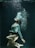 A person underwater, elegantly posed with knees bent and head tilted back. Bubbles rise from their mouth, and they wear a white blouse with black shorts. The water has a deep, dark green hue that contrasts with the lighter tones of their attire.