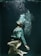 A person underwater, elegantly posed with knees bent and head tilted back. Bubbles rise from their mouth, and they wear a white blouse with black shorts. The water has a deep, dark green hue that contrasts with the lighter tones of their attire.