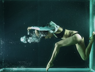 A person is underwater, captured in mid-motion, with bubbles forming around the head and arms. The individual is wearing a black two-piece swimsuit and has limbs gracefully extended. The scene is illuminated by a soft, greenish light, creating an ethereal underwater atmosphere.