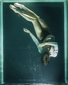 A woman is floating underwater in a glass tank, wearing a light-colored long-sleeve shirt. Her eyes are closed, and her hands are gently placed together in a serene manner. The water is dark and slightly murky, casting an ethereal quality.