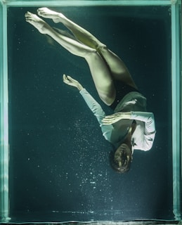 A woman is floating underwater in a glass tank, wearing a light-colored long-sleeve shirt. Her eyes are closed, and her hands are gently placed together in a serene manner. The water is dark and slightly murky, casting an ethereal quality.