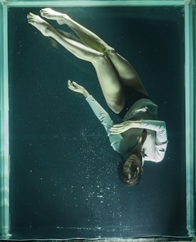 A woman is floating underwater in a glass tank, wearing a light-colored long-sleeve shirt. Her eyes are closed, and her hands are gently placed together in a serene manner. The water is dark and slightly murky, casting an ethereal quality.
