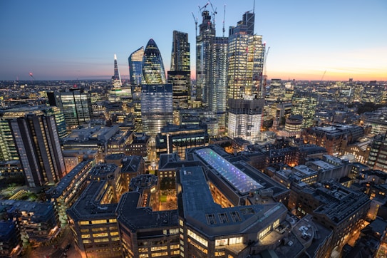 The image captures a bustling cityscape at dusk, featuring a skyline dominated by modern skyscrapers with glass facades reflecting the evening light. The buildings are illuminated with lights, creating a vibrant and dynamic atmosphere. In the background, a blend of contemporary architecture and historical structures can be seen, adding depth to the urban landscape.