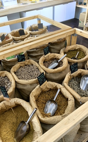 A variety of spices and dry goods are displayed in burlap sacks, each with a scoop, in a well-lit store setting. The bags are organized on wooden shelves, with small blackboards labeling each item. The assortment includes different types of pasta and grains.