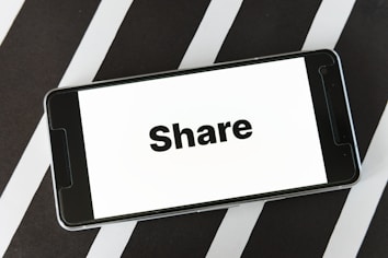 A smartphone lies on a black and white striped surface with the word 'Share' displayed on its screen in bold black letters.