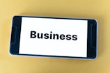 A smartphone lying on a yellow surface displays the word 'Business' in bold black text on its screen.