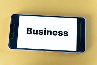 A smartphone lying on a yellow surface displays the word 'Business' in bold black text on its screen.