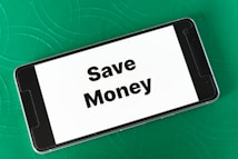 A smartphone displaying the phrase 'Save Money' in bold black letters on a white screen is placed on a green textured surface. The phone is oriented landscape, and the background features subtle circular patterns.