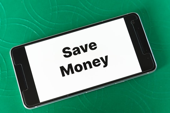 A smartphone displaying the phrase 'Save Money' in bold black letters on a white screen is placed on a green textured surface. The phone is oriented landscape, and the background features subtle circular patterns.