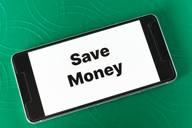 A smartphone displaying the phrase 'Save Money' in bold black letters on a white screen is placed on a green textured surface. The phone is oriented landscape, and the background features subtle circular patterns.