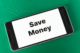 A smartphone displaying the phrase 'Save Money' in bold black letters on a white screen is placed on a green textured surface. The phone is oriented landscape, and the background features subtle circular patterns.